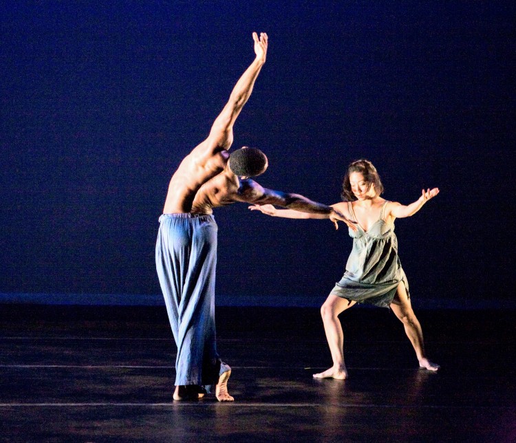 a Black man shirtless in loose light blue pants has his back to us as he bends towards his female partner, an Asian woman in a greenish wrinkled short tunic, she lunges toward him arms out to him, but looks at the floor